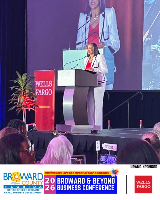 An individual in a stylish suit speaks at a podium with a Wells Fargo banner next to it. The background features a large screen displaying the same person. In the foreground, there are attendees seated. The event is the 2026 Broward & Beyond Business Conference, with Grand Sponsor Wells Fargo.