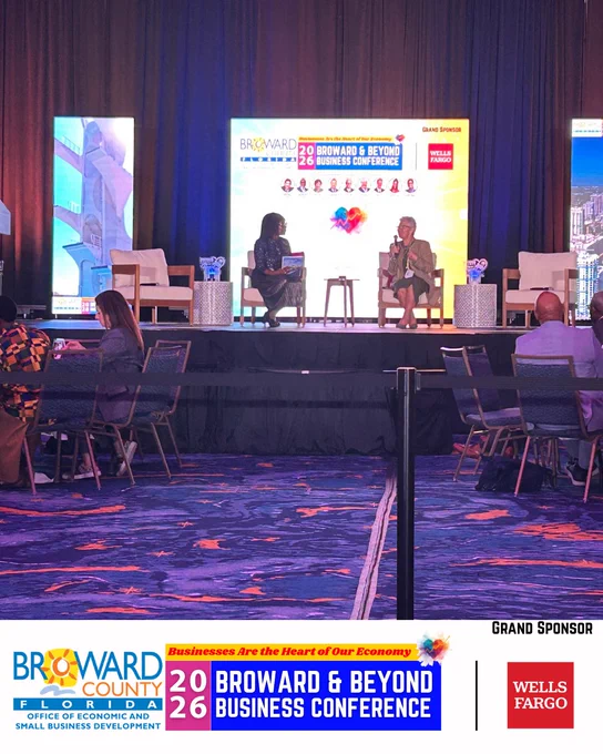 Two individuals engaged in a discussion on stage at the 2026 Broward & Beyond Business Conference. The backdrop displays the event's branding and sponsor logos, including Grand Sponsor Wells Fargo. The stage is equipped with chairs and small tables and the foreground features a colorful carpet.