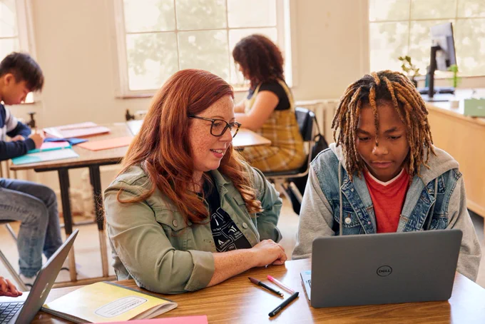 An educator and a student work together at a desk using a laptop in a classroom, with other students studying in the background.