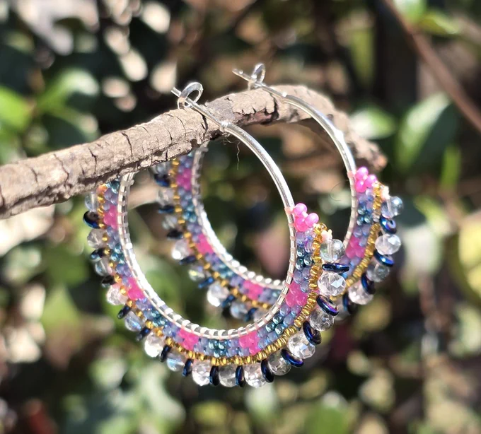 silver-tone hoop earrings with beads in shades of pink, turquoise, iridescent purple, and gold, background of natural wood and greenery 