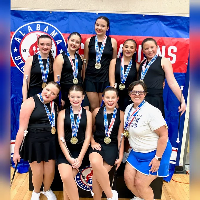 8 girls in black sleeveless dresses in two rows. 5 girls in the back and three in the front. The coach is also in the front row on the left wearing white and blue, school colors. The girls are wearing gold medal necklaces bearing the “Alabama State Games” text and logo. The background is blue with a red stripe that has the Alabama State Games logo with the text “Champions.” 