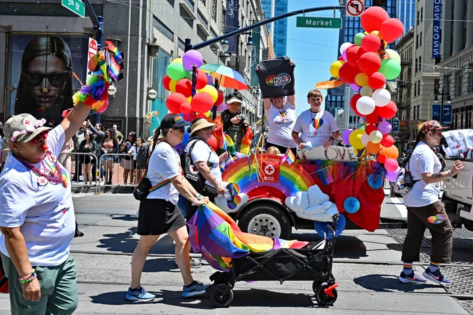 Participants in a vibrant pride parade are walking alongside a colorful float adorned with rainbows, balloons, and umbrellas. The float is on a city street with tall buildings, including a large billboard of Zendaya in the background. Spectators line the sidewalks, enjoying the lively atmosphere.