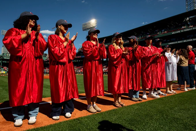 Red Sox Scholars pose on the field smiling and waving wearing graduation gowns and red sox hats.