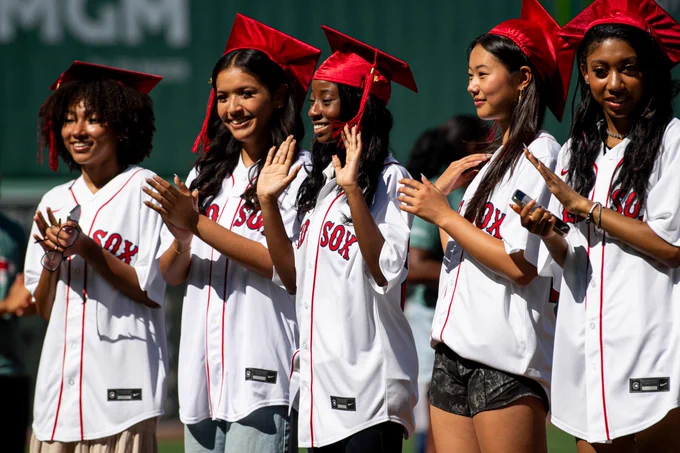 Red Sox Scholars pose on the field smiling and waving wearing white Red Sox uniforms and graduation caps.