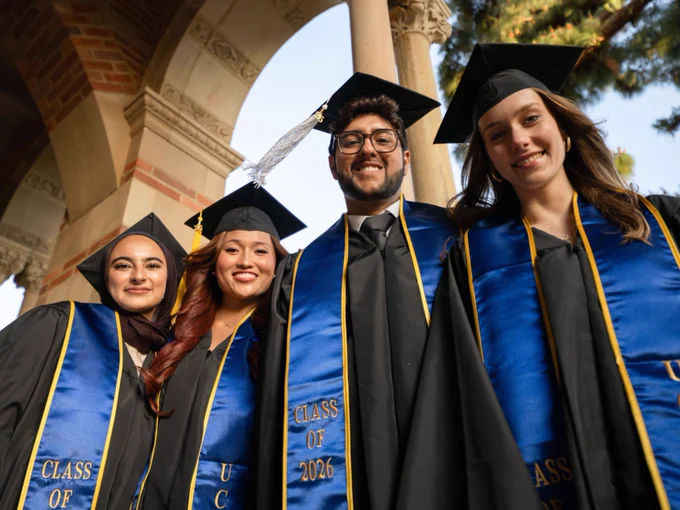 A group of four Bruins wearing caps and gowns stands under an archway at UCLA, with each displaying a blue sash labeled "Class of 2026.