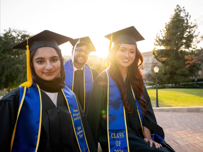 Three UCLA students in graduation attire with blue and gold stoles stand outside. The stoles display "Class of 2026." A sunlit university campus with trees and buildings is in the background.
