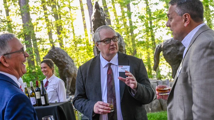 Three men in suits holding drinks and conversing outdoors near a table with wine bottles and a bartender in the background.
