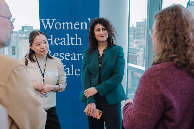 A group of people are gathered in conversation in front of a blue sign that reads "Women's Health Research Yale." Buildings can be seen through large windows in the background.