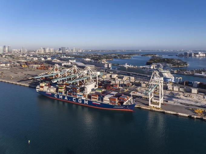 Aerial view of a CMA CGM LNG POWERED SYRACUSE container ship docked at a bustling port. Cranes load/unload colorful containers. In the background, a vibrant cityscape rises, with several large cruise ships nearby, all under a clear blue sky.