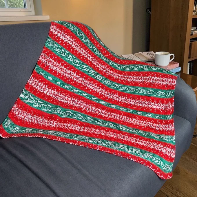 A red, green, and white striped knit blanket lies diagonally across a grey sofa. A small wooden side table holds a white mug on a stack of books and a newspaper. The background features a cream wall, a window, and a wooden bookshelf.