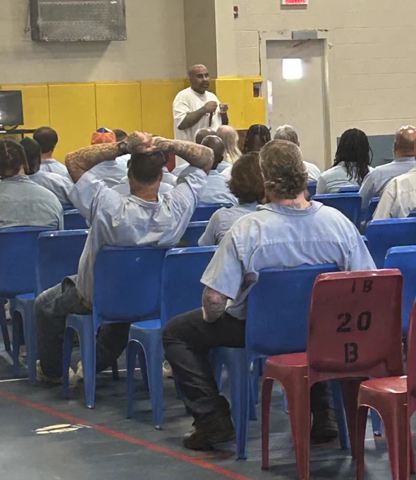 Man with white shirt standing in front of yellow background wall with microphone speaking to seated group with blue collard shirts on in red and blue chairs