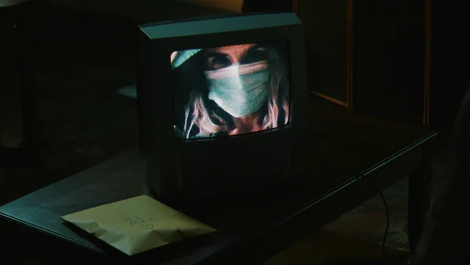 From Cape Fear. A woman appears on a television wearing a face mask and hoodie. Next to the TV is a manila envelope that says, “Hi Max” with a heart underneath.