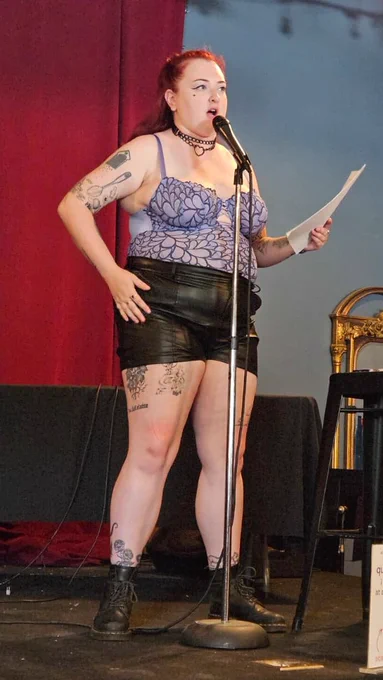 reading a poem at pride while wearing a leather and chain collar, lavender lace bodysuit, and black leather shorts