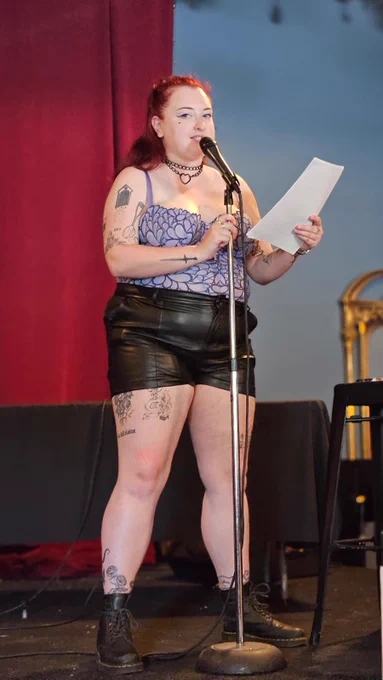 reading a poem at pride while wearing a leather and chain collar, lavender lace bodysuit, and black leather shorts