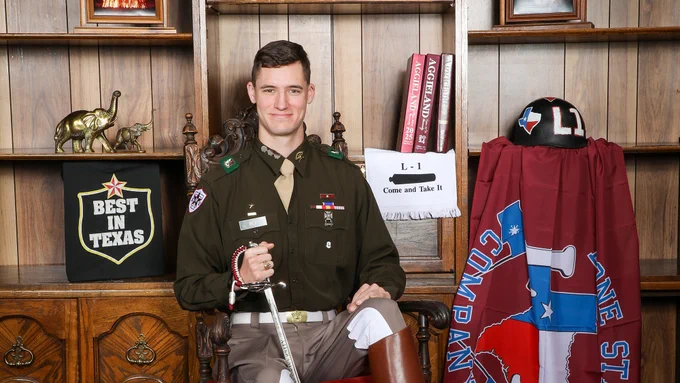 A person in a brown military-style uniform sits in a wooden chair. Nearby, there is a "Best in Texas" sign, a Texas A&M University flag, and a "Come and Take It" towel. A maroon flag with military insignia is draped on a stand.