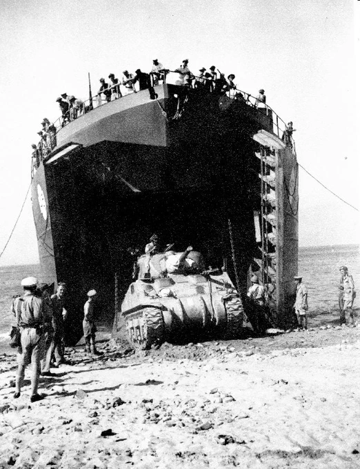 A military tank exits a large landing craft onto a beach. Several people are observing and assisting around the scene. The landing craft is open at the front, with more people visible on top. The setting appears historical with sandy terrain.