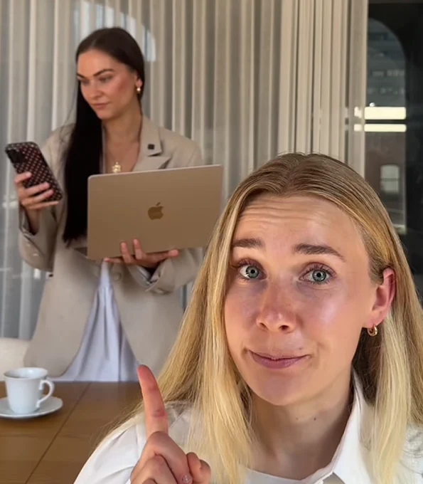 Two women in a modern setting; one focuses on a laptop while the other gestures with a playful expression. A coffee cup is nearby.
