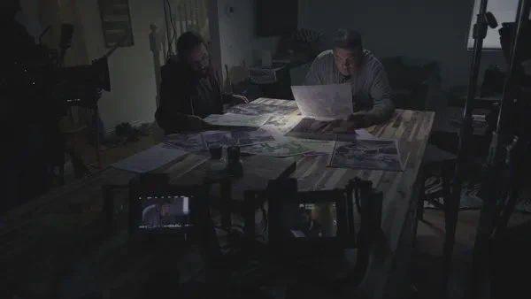 A still from the film Ni-Naadamaadiz: Red Power Rising. Two Native American men sit at a long table, surrounded by film equipment, looking through documents.