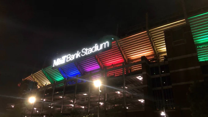 The image shows M&T Bank Stadium illuminated at night with rainbow-colored lights.