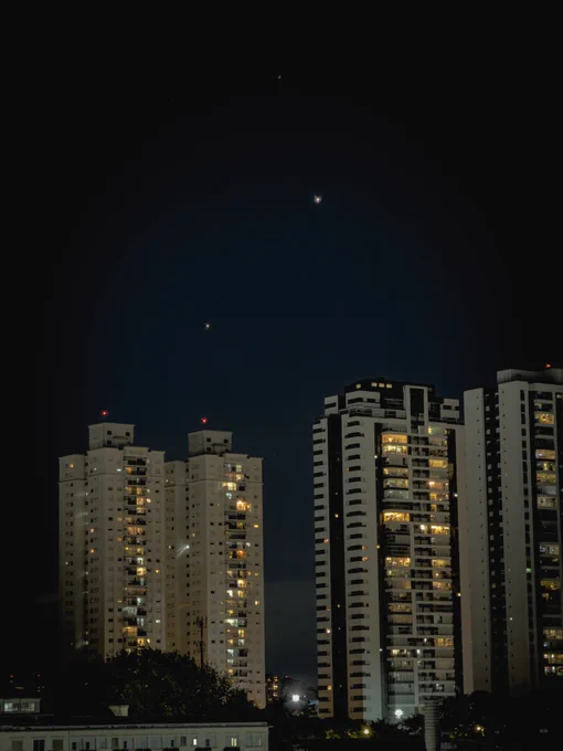 Céu noturno azul profundo sobre São Paulo, com três pontos luminosos visíveis — possivelmente planetas ou estrelas brilhantes em alinhamento diagonal. Prédios residenciais altos dominam o primeiro plano, com janelas iluminadas em tons âmbar revelando a vida urbana noturna. Torres de apartamentos modernos contrastam com edifícios mais antigos à esquerda, todos equipados com luzes vermelhas de sinalização aérea no topo. Vegetação aparece discretamente na base da imagem, entre construções de menor porte. A cena captura a tensão poética entre o cosmos e a metrópole — o céu vasto e silencioso acima de uma cidade que nunca para.
