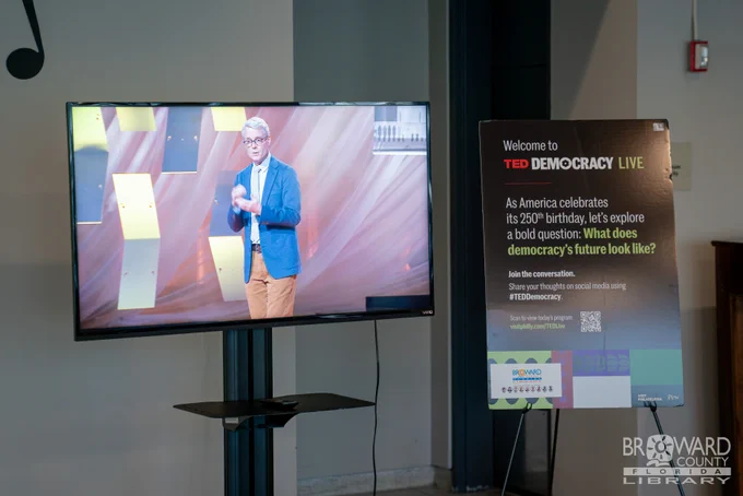 A live stream of a presenter on stage on a television next to TED Democracy signage