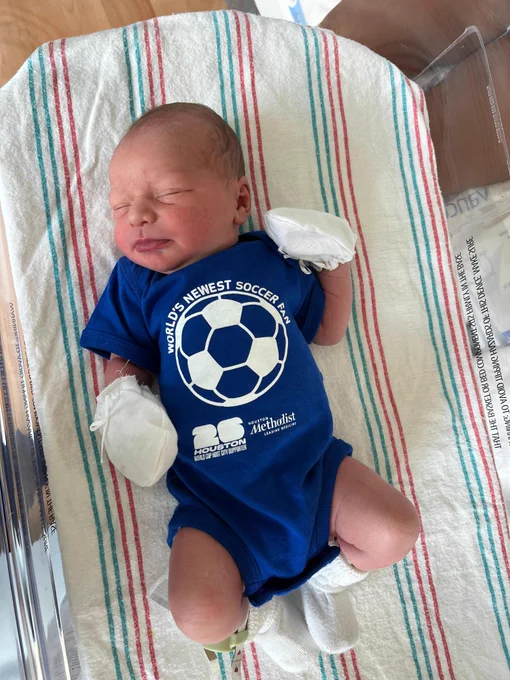 Newborn baby sleeping peacefully on a white, red, and blue striped hospital blanket. The baby wears a blue onesie that reads \"WORLD'S NEWEST SOCCER FAN\" around a soccer ball graphic. Below it are logos for \"Houston 26 World Cup Host City Supporter\" and \"Houston Methodist.\" White mittens and socks are visible.