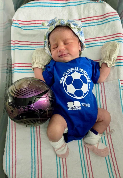 Sleeping newborn in blue onesie with 'WORLD'S NEWEST SOCCER FAN' & a soccer ball graphic. Wearing mittens & socks, lying on a striped blanket. A metallic purple & black soccer ball is next to them. Onesie also reads 'HOUSTON WORLD CUP HOST CITY SUPPORTER' & 'HOUSTON Methodist'.