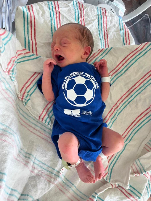 A newborn baby with light skin and dark hair cries while lying on a striped hospital blanket. The baby wears a blue onesie that says \"WORLD'S NEWEST SOCCER FAN\" with a soccer ball. Below it, text reads \"HOUSTON Methodist 20 HOUSTON SUPPORTER\". ID bands are on wrist and ankle.