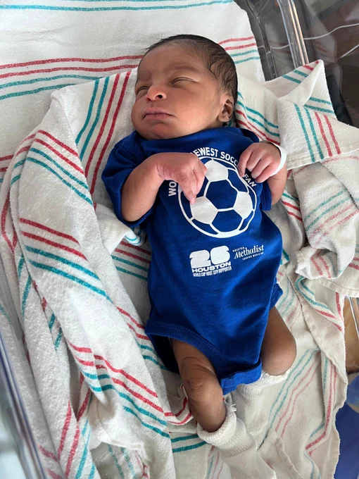 A sleeping newborn baby with dark hair, wearing a blue onesie. The onesie features a soccer ball graphic and text: \"WORLD'S NEWEST SOCCER FAN\", \"HOUSTON 26 WORLD CUP HOST CITY SUPPORTER\", and \"Houston Methodist LEADING MEDICINE\". The baby is on a white blanket with red and blue stripes.
