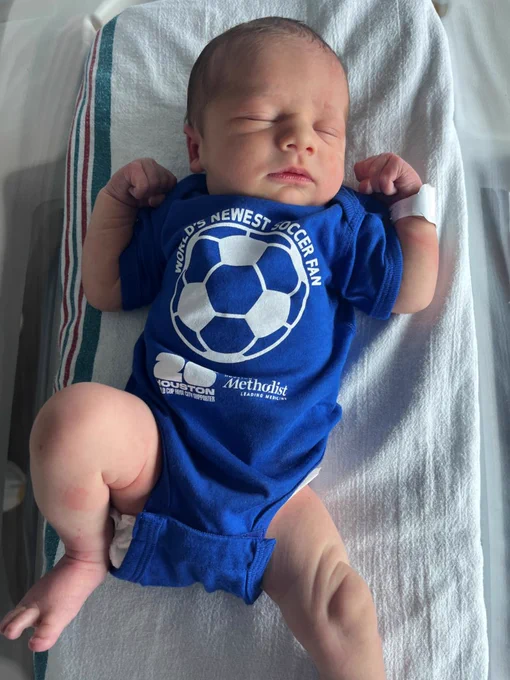 Newborn baby with light skin, eyes closed, sleeping on a hospital blanket. Wears a blue onesie with \"WORLD'S NEWEST SOCCER FAN\" around a soccer ball, plus \"HOUSTON WORLD CUP HOST CITY SUPPORTER\" and \"Methodist LEADING MEDICINE\".