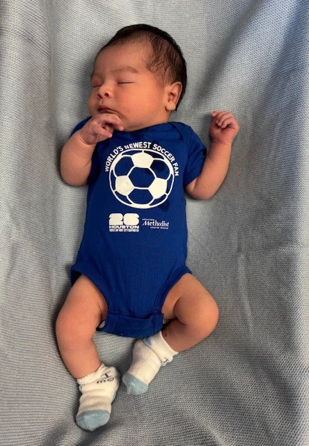 Newborn baby sleeping peacefully on a light blue blanket. The baby wears a blue onesie with \"WORLD'S NEWEST SOCCER FAN\" and a soccer ball logo. Below it, \"26 HOUSTON Methodist LEADING MEDICINE\". White socks with light blue toes are on its feet, one sock says \"1 mo\".