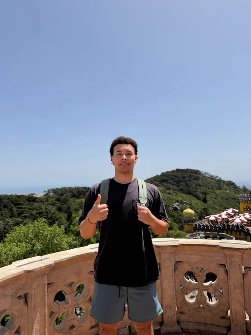 A person standing near lush green hills and a bright blue sky, with an ornate structure partially visible, resembling architectural designs from places like the Pena Palace in Sintra, Portugal.