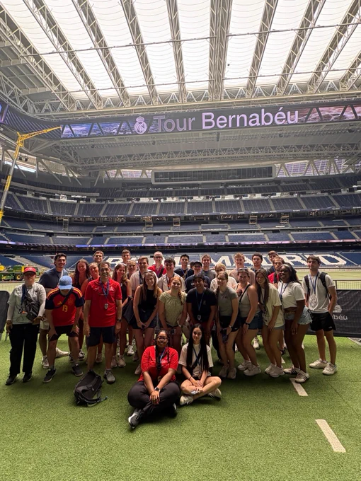 A group of people are posing for a photo inside Santiago Bernabéu Stadium. They are standing on the grass with the stadium seating visible behind them. A large screen above displays "Tour Bernabéu.