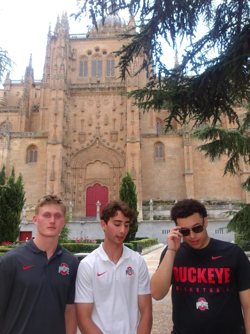 Three individuals stand in front of the historic facade of a large cathedral, featuring intricate architectural details. Two of them are wearing Ohio State University shirts, and one is adjusting sunglasses. Trees and plants are visible in the foreground.
