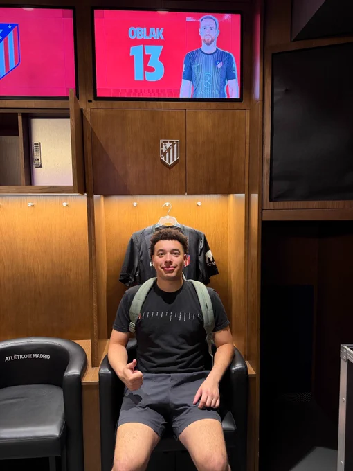 A person sitting in a locker room chair with a backpack, giving a thumbs-up. The background shows a screen displaying an image of a player with the name "Oblak" and the number 13, along with Atlético de Madrid branding.