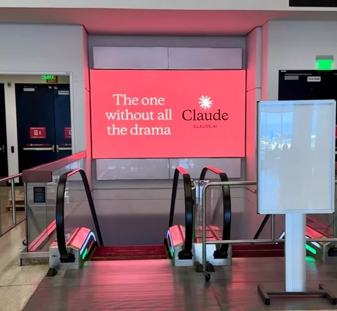 A vibrant digital display features text promoting "Claude" as a solution without drama, positioned near escalators in a public space.