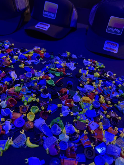 A vibrant collection of assorted LEGO minifigure parts scattered on a table under blue lighting, with black caps labeled 'mighty' in the background. The scene has a playful and creative mood.