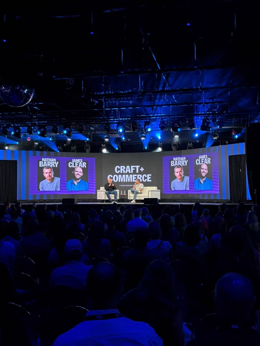 Wide-angle view of a stage at the Craft   Commerce conference with two speakers in conversation under blue lighting. The audience is engaged and seated in rows.
