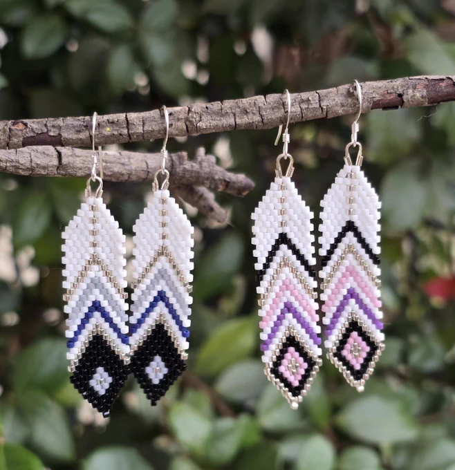 beaded feather earrings with white background, one pair has shades of blue, gray, black, and silver, the other has shades of black, lavender, pink, and silver, with .925 sterling silver ear hooks, background of natural greenery 
