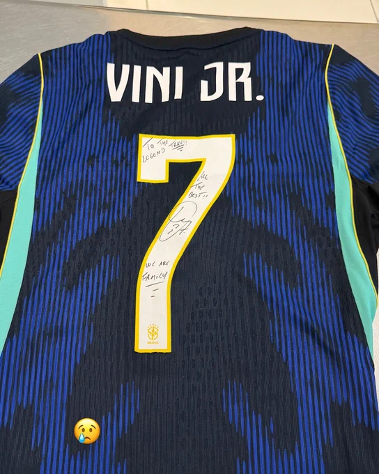 A Vini Jr. football jersey with a handwritten message and signature. The jersey features the name and number 7 on the back, with a crying emoji at the bottom.