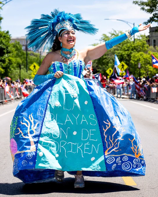 3. A Puerto Rican Chicagoan woman wearing an elaborate light blue structured dress and feathered headpiece, saying 'La Playas Son de Boriken', marches at the Puerto Rican People's Day Parade in Humboldt Park.