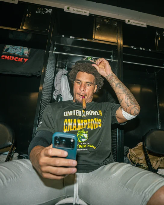 A football player celebrates a championship victory in the locker room, sitting with a cigar and taking a selfie. The mood is joyful and relaxed post-game.
