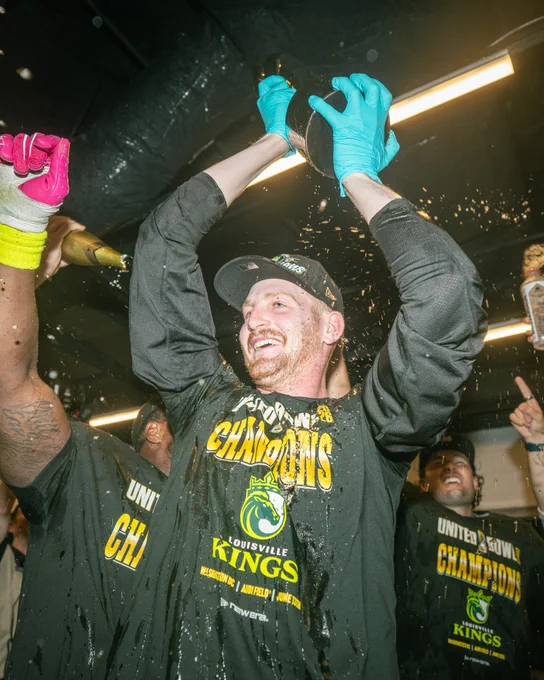 Team members celebrate their victory with champagne, wearing championship shirts. Excitement and jubilation fill the room.