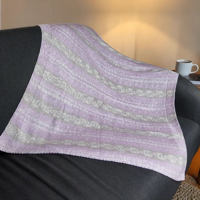 A hand-knitted lilac and grey striped blanket rests on a dark grey sofa. The blanket features a seed stitch border. In the background, a white mug sits on a table beside a warm lamp, creating a cosy atmosphere in a dimly lit room.