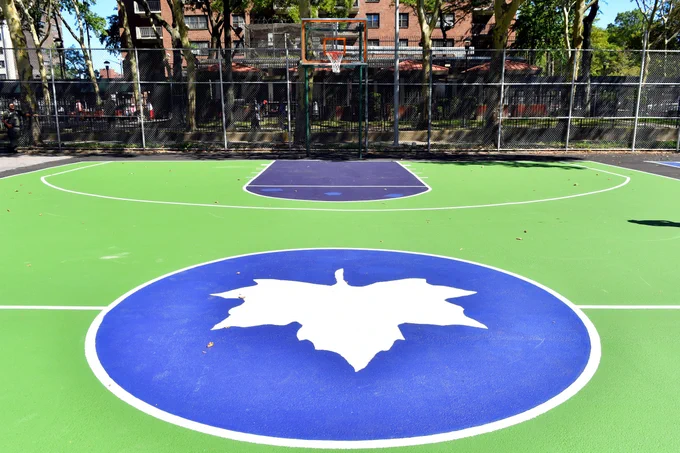 A city basketball court, with the iconic park leaf in the middle of the photo. The court is brightly painted, and familiar NYC-style housing complexes can be seen in the distance.