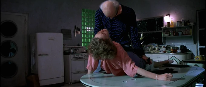 Manhunter screenshot of Tom Noonan as Francis Dollarhyde and Joan Allen as Reba in Dollarhyde's kitchen. the pair are slightly to the right of frame/off-center, Dollarhyde stands, wearing a blouse with a pattern like an EKG machine reading, little squiggly lines of cobalt blue against black. he holds a mirror shard in his left hand. Reba is lying supine on the kitchen table, partially lifted up so her face is angled toward Dollarhyde's, blonde hair falling behind her. his kitchen is a mess but it has some cool emerald green glass bricks right behind him, and a mint green table with incredible art deco-like chairs with a diamond-shaped back 