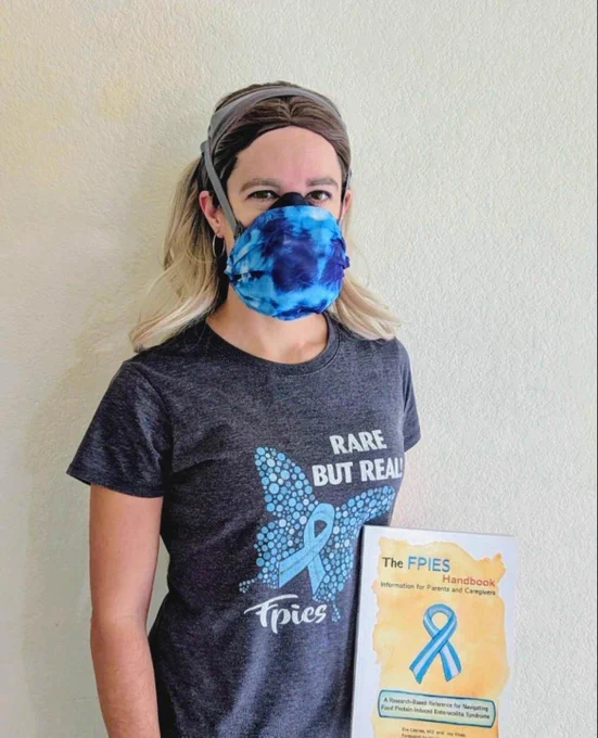 A Caucasian woman with blonde hair is wearing a GVS Elipse with a blue and black tie dye cloth cover, and a shirt that says Rare but Real: FPIES. She is holding a book that says "The FPIES Handbook" and has a blue ribbon drawn on the cover. 