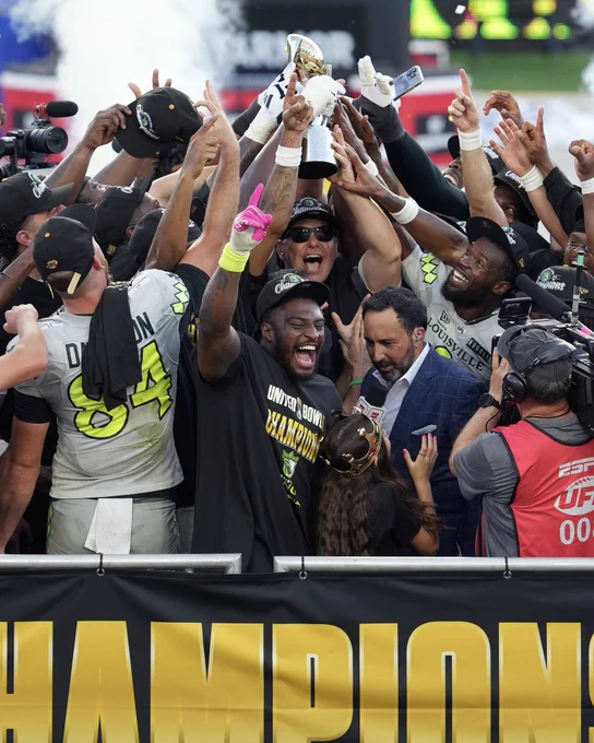 A joyous celebration with a sports team holding a trophy. The players and staff are gathered in excitement, wearing championship apparel.