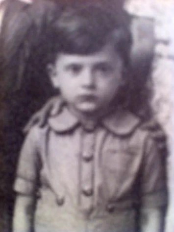 Black and white portrait of a young boy standing and looking directly at the camera, wearing a buttoned shirt with a distinctive collar. 
