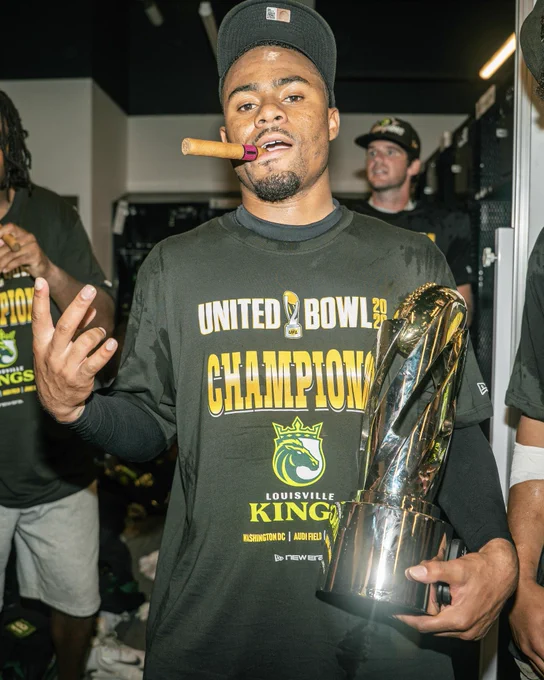 A triumphant football player celebrates holding a shiny trophy and a cigar in a locker room. He wears a championship t-shirt with the text 'United Bowl Champions' and 'Louisville Kings'. The mood is celebratory and victorious.